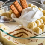 Banana pudding in a glass dish, showcasing layers of custard, bananas, and vanilla wafers, topped with whipped cream and garnished.